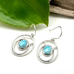 K&ouml;nnte beinhalten: Ein Paar silberne Ohrringe mit einem hellblauen Edelstein, der in einem runden Rahmen gefasst ist. Die Ohrringe haben ein schlichtes, elegantes Design.