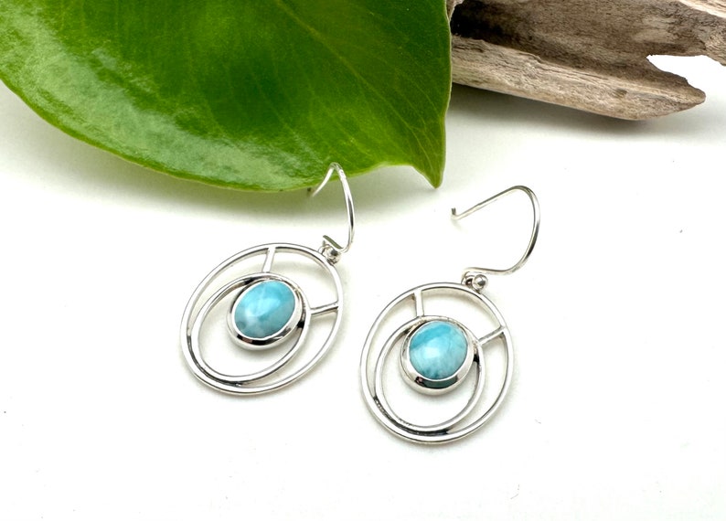 K&ouml;nnte beinhalten: Ein Paar silberne Ohrringe mit einem hellblauen Edelstein, der in einem runden Rahmen gefasst ist. Die Ohrringe haben ein schlichtes, elegantes Design.