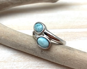 May include: A silver-colored ring with two oval, light blue gemstones. The ring's band curves around the finger, with the gemstones set in a bezel style. The ring is displayed on a piece of driftwood.