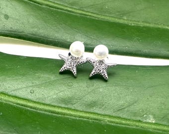 Puede incluir: Un par de pendientes de estrella de mar plateados con detalles de perlas blancas. Los pendientes están sobre un fondo de hoja verde.