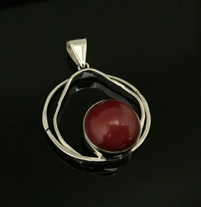 Pu&ograve; includere: Un ciondolo d'argento con una pietra preziosa rossa incastonata in un design a doppio cerchio.