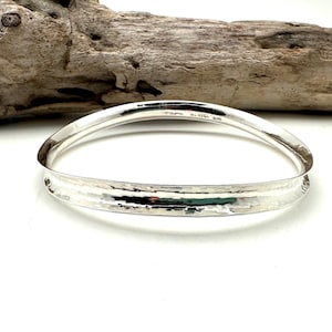 May include: A silver hammered bangle bracelet with a wide, rounded band. The bracelet is smooth and polished, with a hammered texture on the front.