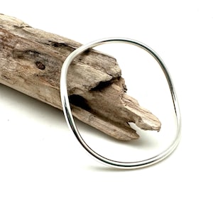 May include: A simple silver bangle bracelet, shown resting on a piece of driftwood.