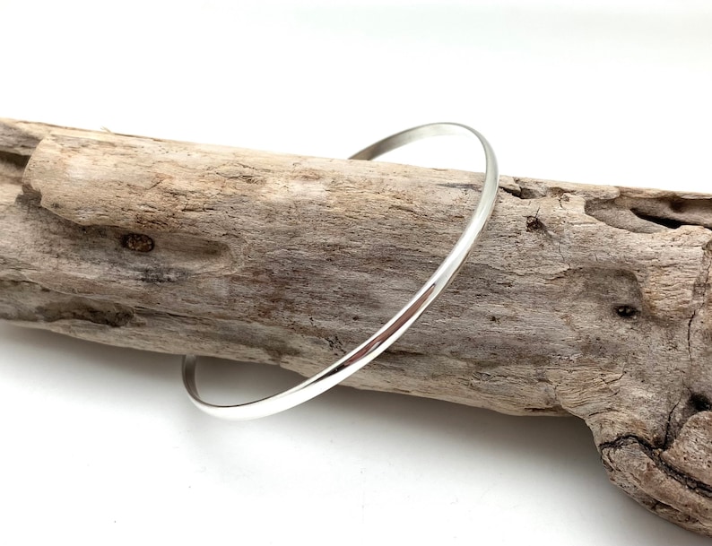 May include: A simple silver bangle bracelet, resting on a piece of driftwood.