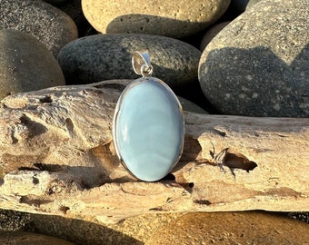May include: A silver oval pendant with a light blue gemstone. The pendant is sitting on a piece of driftwood.