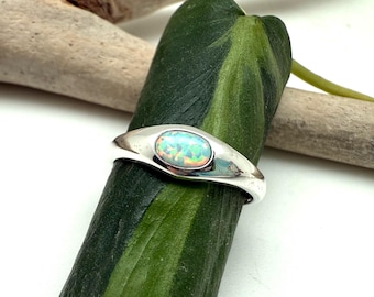 May include: A silver ring with an oval opal gemstone. The opal displays a vibrant play of color, including shades of blue, green, and orange. The ring is set against a green leaf and a piece of driftwood.