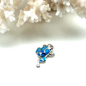 Puede incluir: Un amuleto de rana de plata con incrustaciones de ópalo azul. La rana está sentada sobre una superficie blanca.