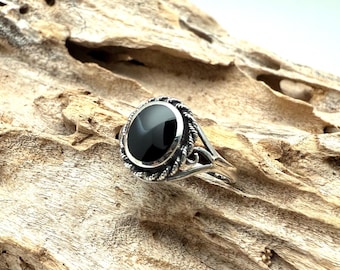 May include: A silver ring featuring an oval, black gemstone. The ring has a decorative, twisted rope-like border around the stone and ornate detailing on the band. The ring is set against a textured, light brown wooden surface.