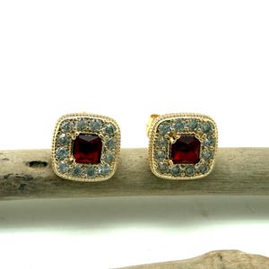 May include: A pair of gold-toned square stud earrings. Each earring features a central, square, deep red gemstone surrounded by a halo of smaller, clear gemstones. The earrings are set on a wooden surface against a white background.