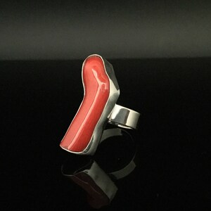 May include: A silver ring with a large, bright red coral stone set in the band. The stone is shaped like a long, thin, irregular shape.