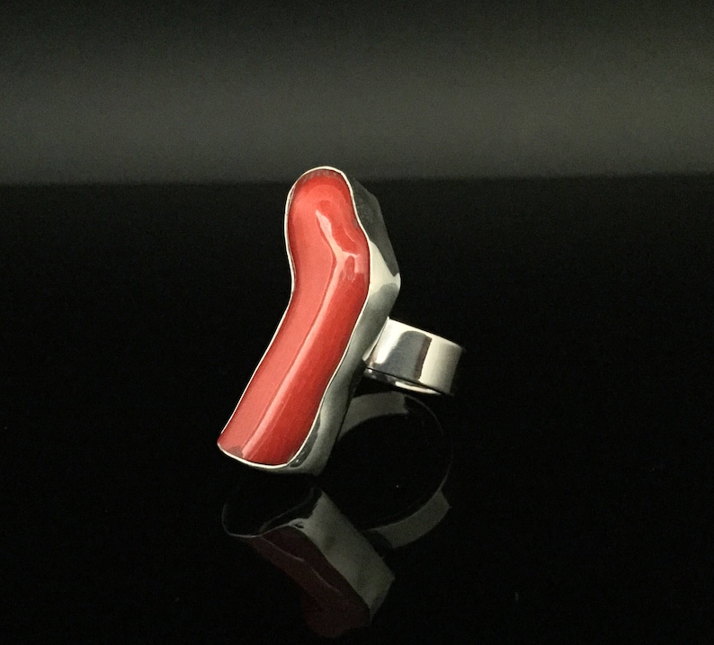 May include: A silver ring with a large, bright red coral stone set in the band. The stone is shaped like a long, thin, irregular shape.