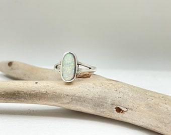 May include: A silver ring with an oval-shaped white opal gemstone. The ring has a delicate design with two prongs on each side of the stone.
