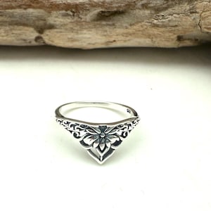 May include: A silver ring with an ornate floral design. The ring features a central flower motif with detailed petal work, set within a decorative, openwork band. The ring is displayed on a white surface, with a wooden element in the background.