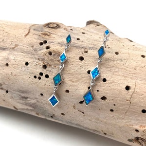 Op de afbeelding: Een paar zilveren oorhangers met ruitvormige blauwe opaalstenen. De oorbellen worden gepresenteerd op een stuk drijfhout, wat hun ontwerp en kleur benadrukt. Elke oorbel heeft meerdere opaalstenen.