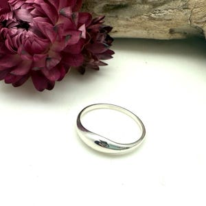 Puede incluir: Un anillo de plata pulida con un diseño suave y redondeado. El anillo tiene forma de lágrima en un lado. Una flor de color burdeos oscuro y un trozo de madera flotante están en el fondo, sobre una superficie blanca.