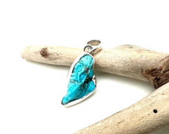 May include: A turquoise pendant with a silver setting. The pendant is shaped like a teardrop and has a rough, natural texture. It is resting on a piece of driftwood.