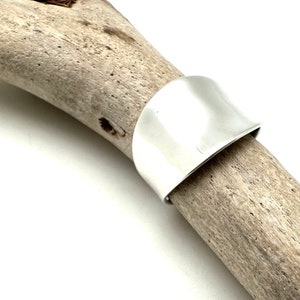 May include: A silver ring with a wide, flat band. The ring is smooth and polished, and it has a slightly curved shape.