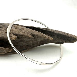 May include: A simple silver bangle bracelet with a slightly curved shape. The bracelet is resting on a piece of driftwood.