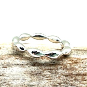 May include: A silver ring with a repeating oval design. The ring is made of a shiny, reflective metal and sits on a piece of weathered wood. The ring's design is a series of connected, elongated ovals.