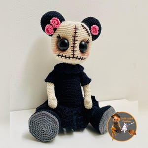May include: Handmade crochet doll with a stitched face, large black eyes, and pink flower accents on the ears. The doll is dressed in a black outfit and has grey feet. The doll has a unique, whimsical design.