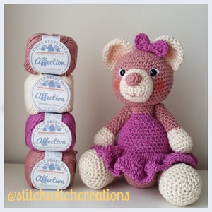 Könnte beinhalten: Drei Knäuel rosa, weiß und beigefarbenes Garn mit der Aufschrift "Three Bears Yarn Affection 100% Aras Cotton" sind übereinander gestapelt. Ein gehäkelter rosa Bär mit einem lila Kleid sitzt vor dem Garn.