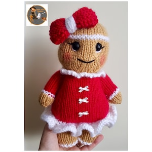 Puede incluir: Una muñeca de jengibre de punto que lleva un vestido rojo con ribetes blancos y un lazo rojo. La muñeca tiene una cara sonriente con ojos negros y mejillas sonrosadas.