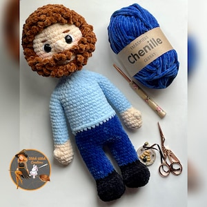 May include: A handmade crochet doll with a brown beard and hair, wearing a light blue shirt and dark blue trousers. A ball of blue chenille yarn, a crochet hook, and scissors are also present. The doll is approximately 30 cm tall.