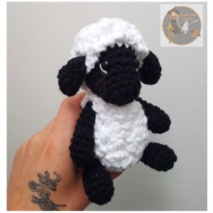 May include: A crocheted black and white sheep plush toy. The sheep has a white body and a black face, legs, and ears. The sheep is sitting on a hand.