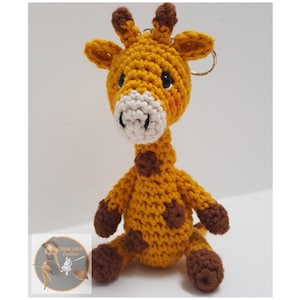 Könnte beinhalten: Ein gehäkelter Giraffen-Schlüsselanhänger mit braunen Flecken, einem weißen Gesicht und braunen Beinen. Die Giraffe sitzt auf einer weißen Oberfläche.