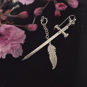 May include: Silver sword earring with a feather charm hanging from a chain. The earring is on a black background with pink flowers.