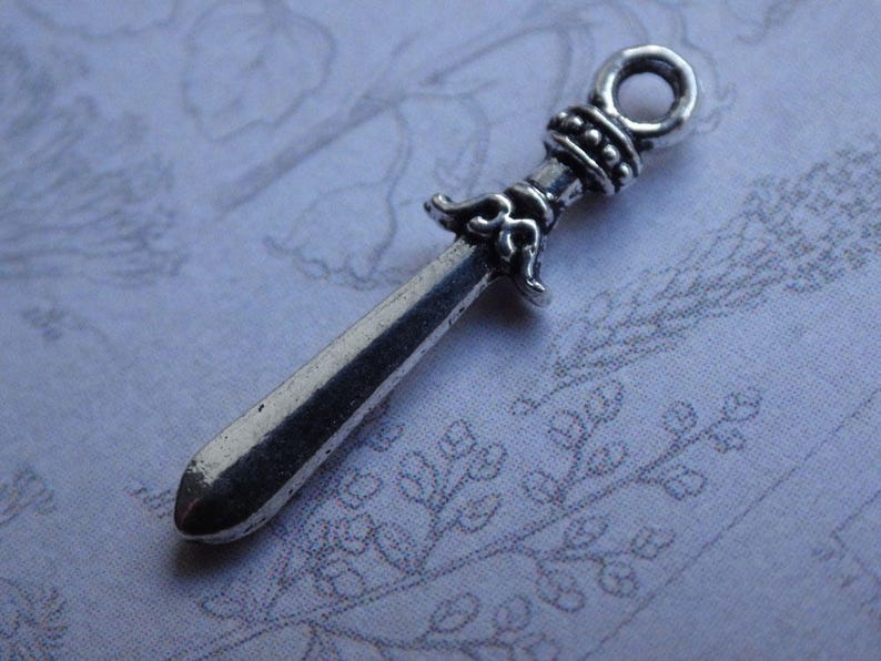 Puede incluir: Encanto de plata en forma de espada con un mango adornado. La espada mide aproximadamente 5 cm de largo.