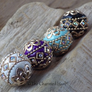 Peut inclure: Quatre perles rondes décoratives avec des filigranes dorés et des strass. Les perles sont blanches, violettes, turquoise et noires.