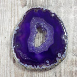 May include: A polished, oval-shaped purple agate geode slice. The geode features a deep purple center with a crystalline cavity and a lighter purple outer band. The outer edge has a white and tan speckled pattern.