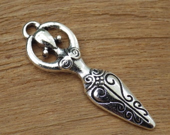 May include: A silver-colored pendant with an ornate design. The pendant features a loop at the top, a central figure with a circular element, and a stylized, elongated body with decorative swirls and patterns. The pendant is on a wooden surface.
