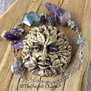 May include: A pendant featuring a gold-coloured Green Man face with leaf details, amethyst stones, and a purple crystal pendulum. The pendant is on a silver chain and rests on a wooden surface.