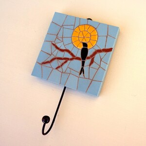 May include: A blue mosaic tile wall hanging with a black bird silhouette perched on a brown branch. The bird is facing a yellow sun-like design. The wall hanging has a black metal hook.