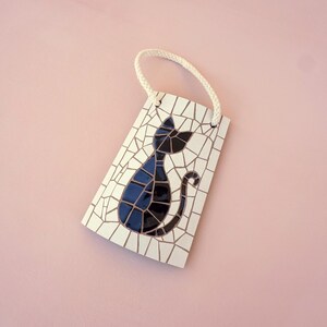 May include: A white ceramic tile with a black mosaic cat design. The tile has a white rope hanger attached to the top.