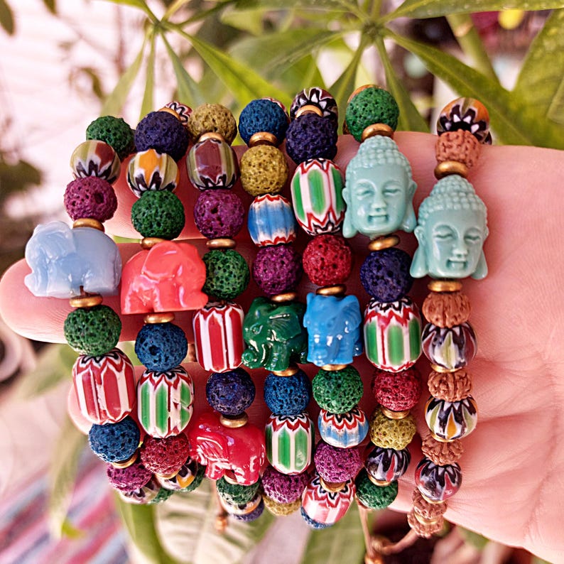 Pu&ograve; includere: Un set di braccialetti colorati con perline di roccia vulcanica, piccoli ciondoli a forma di elefante e ciondoli a forma di testa di Buddha. I braccialetti sono impilati su una mano.