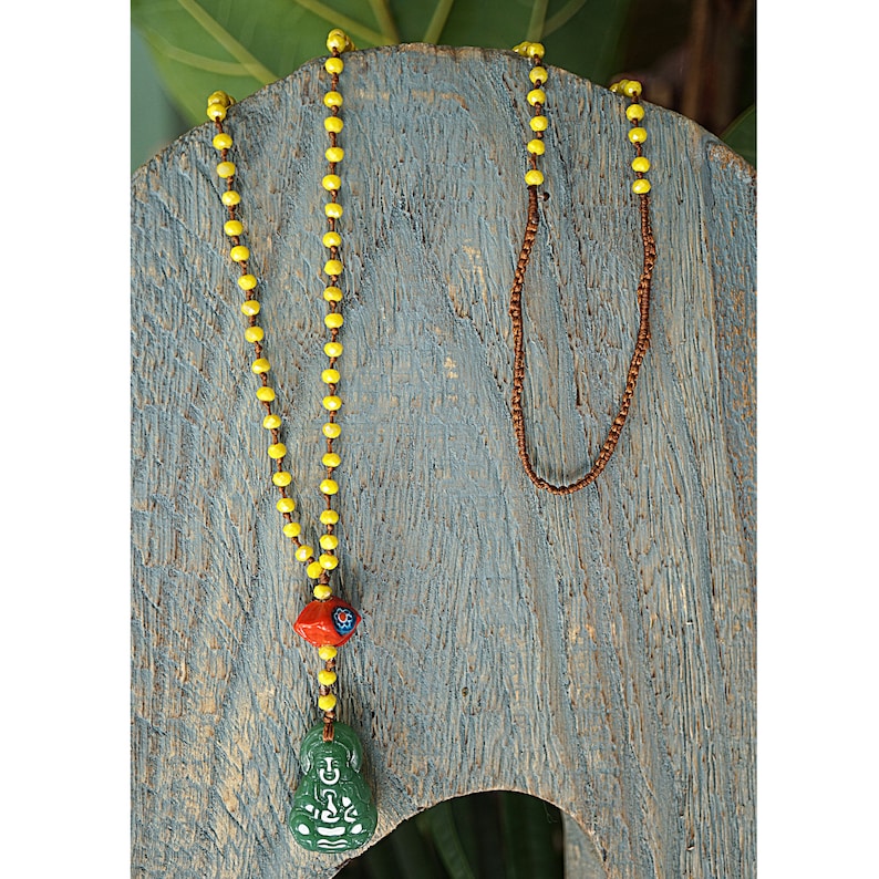 May include: A yellow beaded necklace with a green jade Buddha pendant and a red and blue bead accent. The necklace is displayed on a wooden surface.