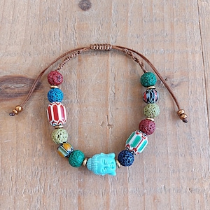 Pu&ograve; includere: Un braccialetto multi-colorato con un ciondolo a forma di Buddha blu. Il braccialetto presenta perline di roccia lavica, perline di ceramica con motivi e un cordoncino marrone.