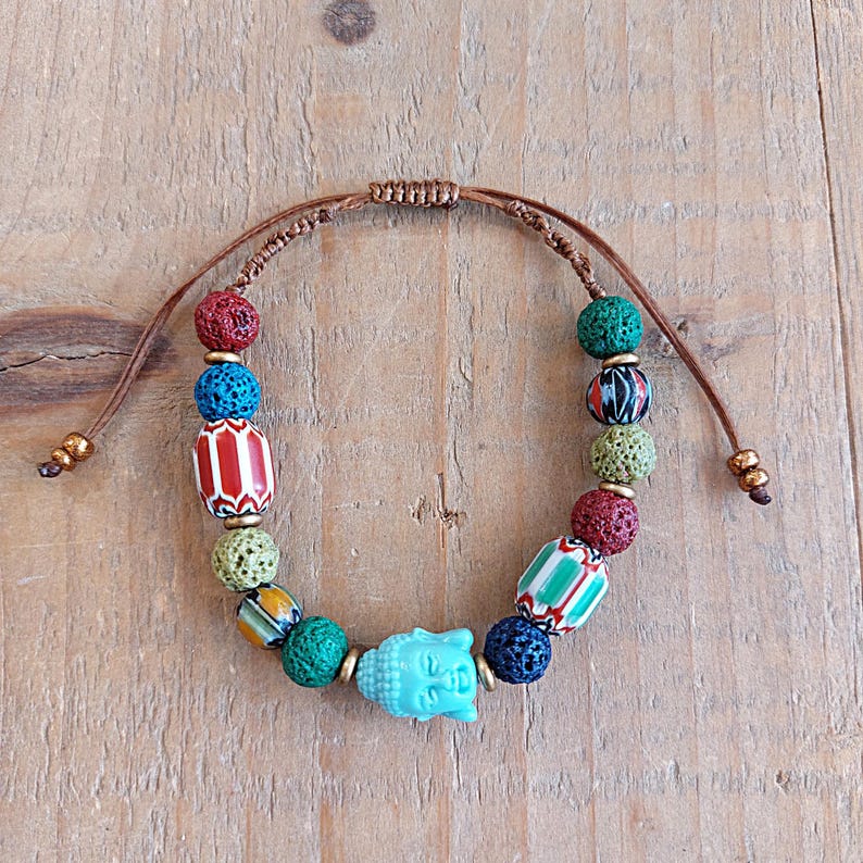 Pu&ograve; includere: Un braccialetto multi-colorato con un ciondolo a forma di Buddha blu. Il braccialetto presenta perline di roccia lavica, perline di ceramica con motivi e un cordoncino marrone.