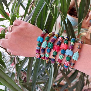 Pu&ograve; includere: Sette braccialetti colorati con perline di roccia vulcanica e piccoli ciondoli di elefante e Buddha in ceramica. I braccialetti sono impilati sul polso di una persona.