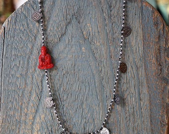 May include: A silver chain necklace with small round beads and silver coin charms. A red figurine of a seated Buddha is attached to the chain.