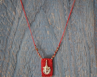 May include: A red macrame necklace with a gold-colored pendant. The pendant features an ornate design. The necklace has a red cord and gold-colored beads. The pendant is approximately 1 inch in length.