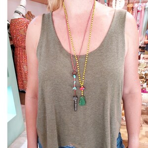 May include: A woman wearing a green tank top with three necklaces. The necklaces are made of different materials and have various charms, including a green Buddha pendant, a red and turquoise charm, and a long, brown charm.