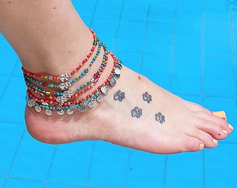 May include: A woman's ankle with a layered anklet made of colourful beads and silver charms. The anklet is adorned with small silver charms shaped like hands. The ankle has a tattoo of four paw prints.