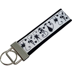 May include: A black and white cat-themed keychain wristlet. The white ribbon features black cat silhouettes, hearts, and swirls. The wristlet is attached to a silver-colored metal key ring.