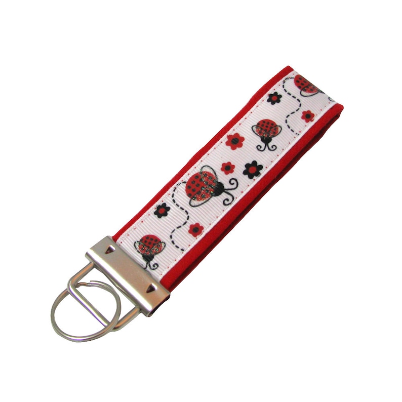 Puede incluir: Llavero rojo y blanco con una mariquita y un anillo de llave plateado. El llavero tiene un respaldo de tela roja y un frente de tela blanca con un patr&oacute;n repetido de mariquitas y flores negras.