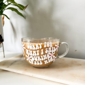 Op de afbeelding: Een heldere glazen mok met een witte handgreep gevuld met koffie. De mok heeft een wit ontwerp met roze bloemen en de tekst "Sorry for what I said during hell week".