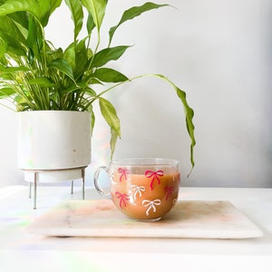 May include: A white ceramic mug with a pink and white bow pattern filled with a brown beverage. The mug is sitting on a white marble chopping board.